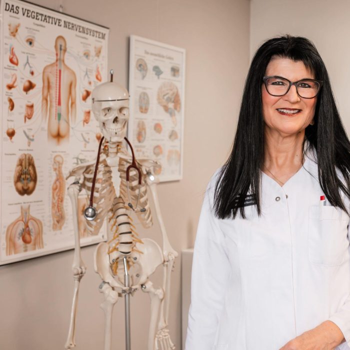 Dr. med. Sabine Ott-Oechsle steht neben einem Skelettmodell vor einer Infotafel zum vegetativen Nervensystem in ihrer Privatpraxis in Illertissen.